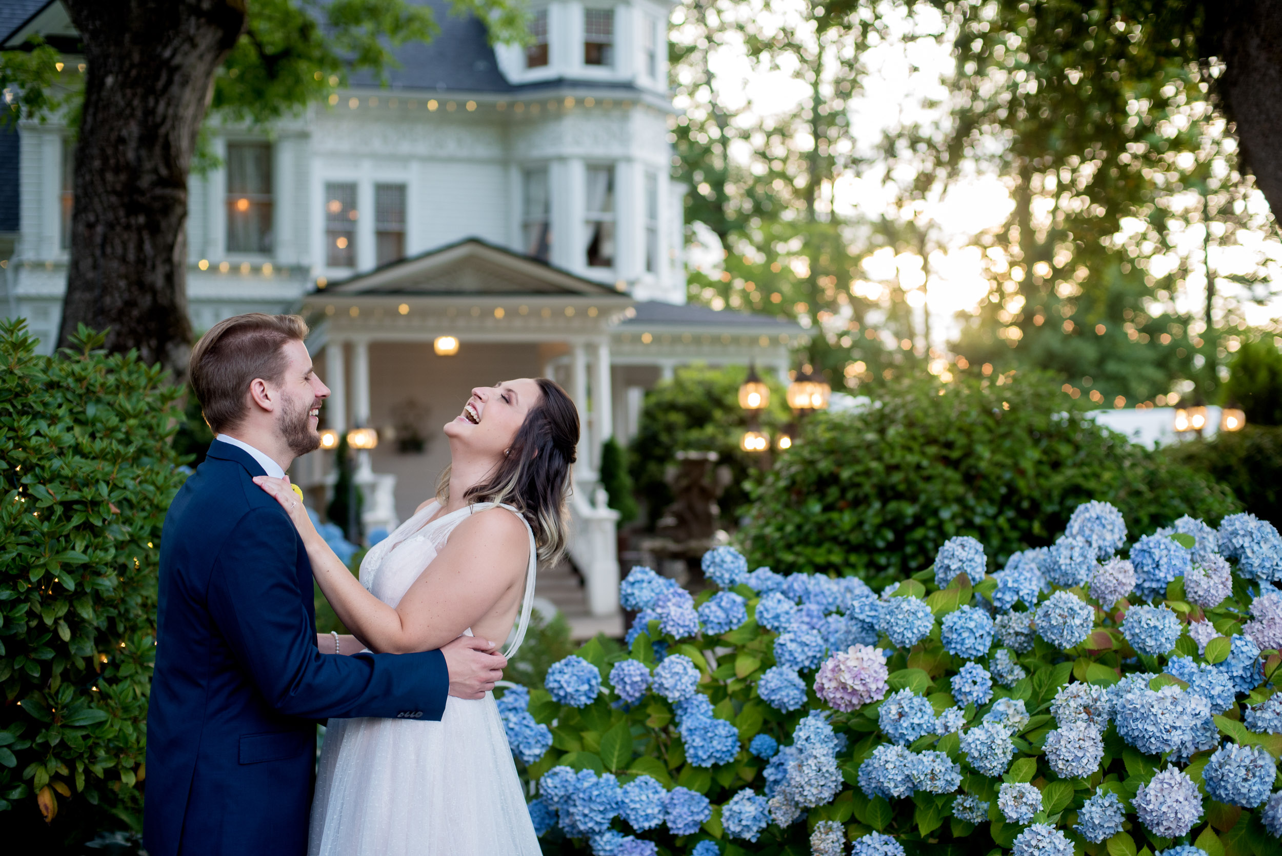 a bride and groom laugh standing in front of hydrangeas with victorian belle mansion in background