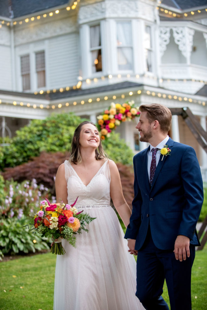 a bride and groom walk together holding colorful yellow orange and pink bouquet while standing in front of the garden at victorian belle mansion with lights in the background