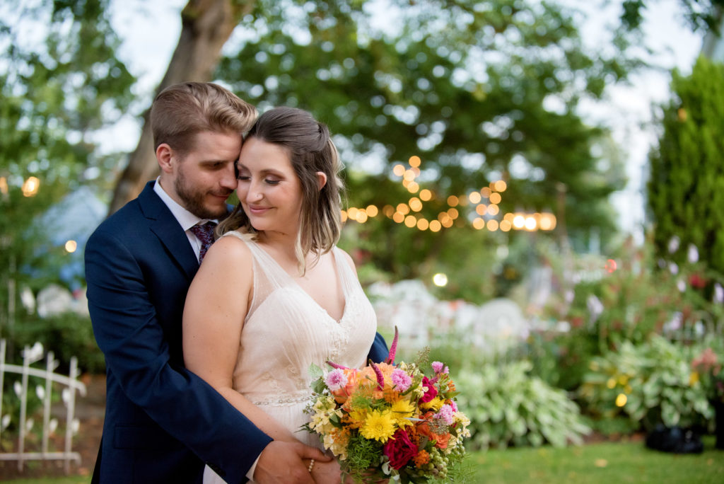 a bride and groom hug together holding colorful yellow orange and pink bouquet while standing in front of the garden at victorian belle mansion with lights in the background