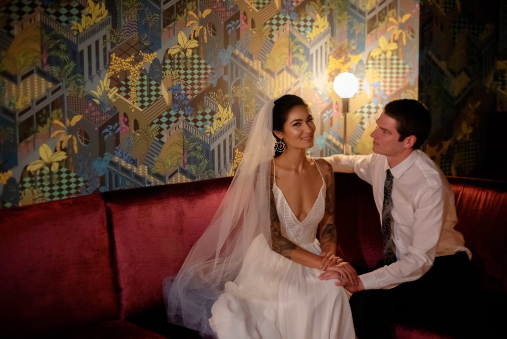 bride and groom sit on the velvet couch under colorful wallpaper at voysey speakeasy bar under The Evergreen